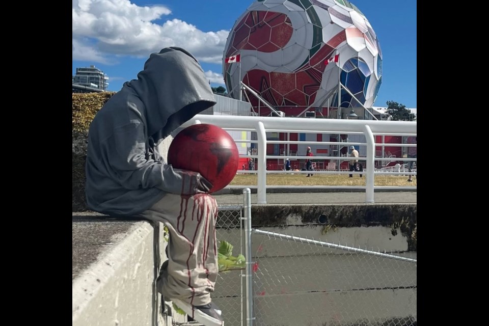 A hooded child sculpture cradling a blood-red globe, with red running down its legs, sitting on a wall above the water in front of Science World's geodesic dome wrapped as a giant FIFA World Cup match ball.
